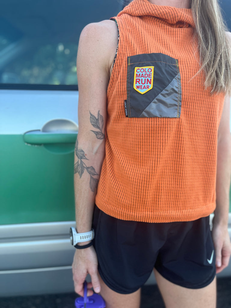 Person wearing an orange sleeveless jacket with a visible brand logo, standing in front of a green car.
