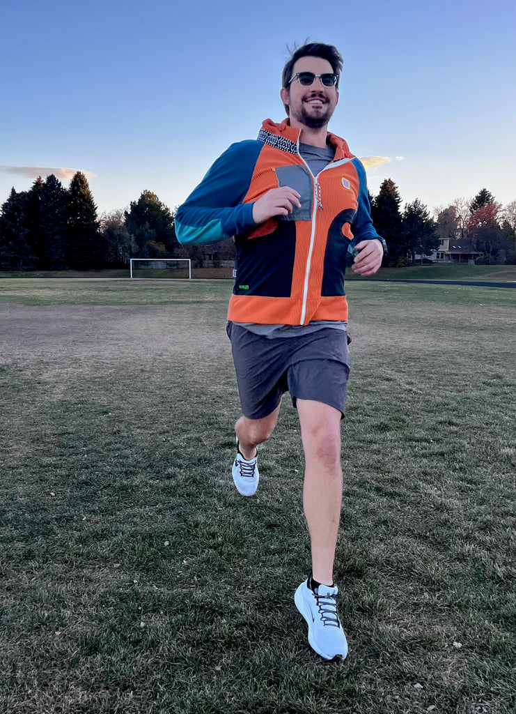 Man running on a grassy field wearing an orange and blue jacket.